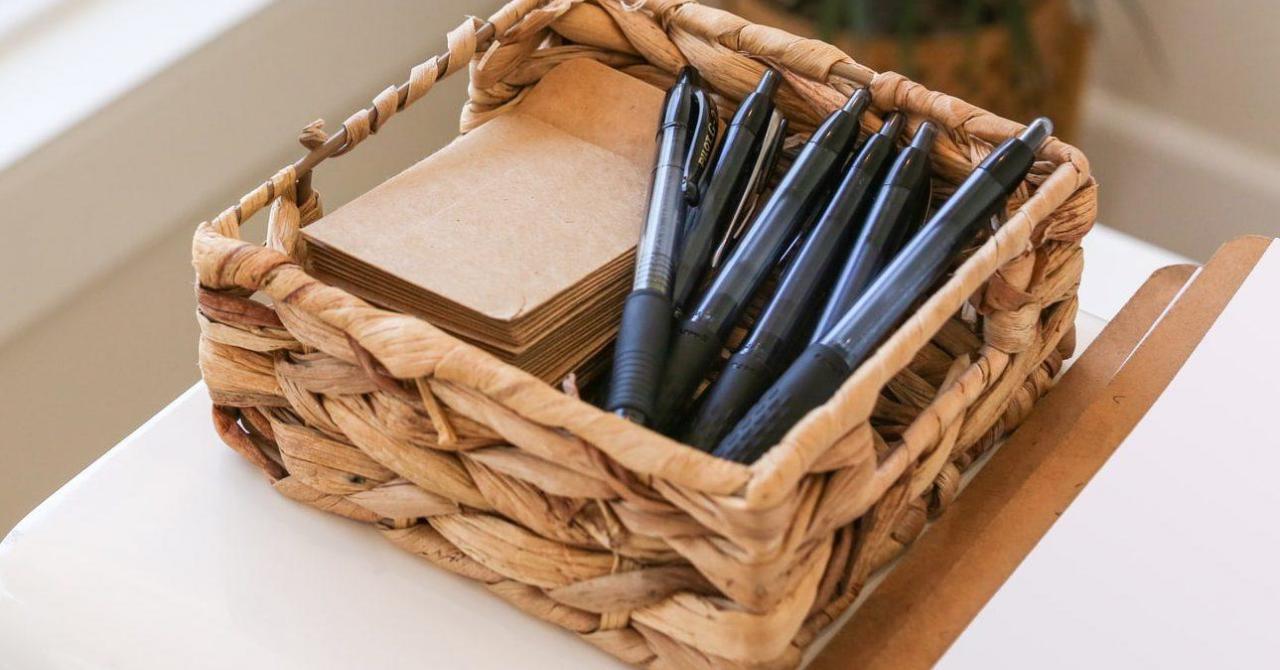 A small, square rattan basket holds a stack of brown paper, and a collection of pens