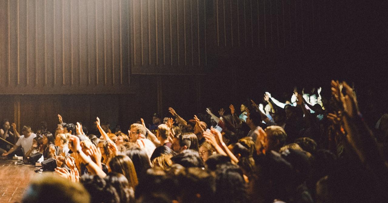 A crowd of people in a large auditorium are raising their hands. The auditorium has light streaming in from above.