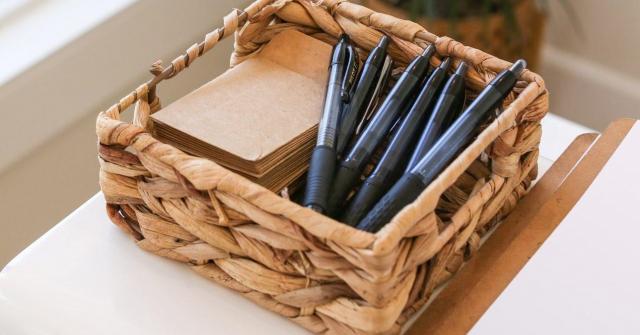 A small, square rattan basket holds a stack of brown paper, and a collection of pens